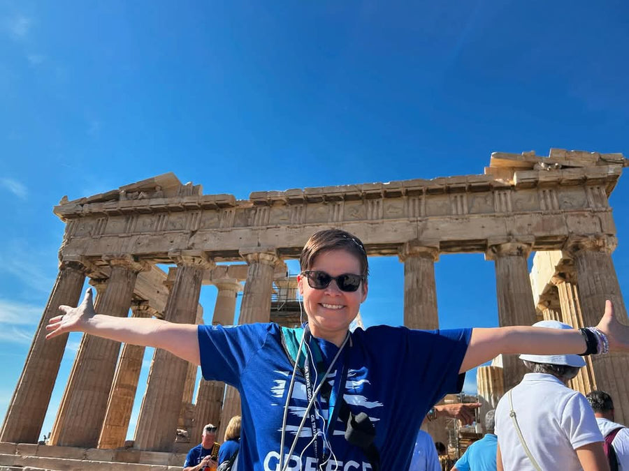 Elisabeth in a blue shirt with arms outstretched in front of the Parthenon against a clear blue sky.